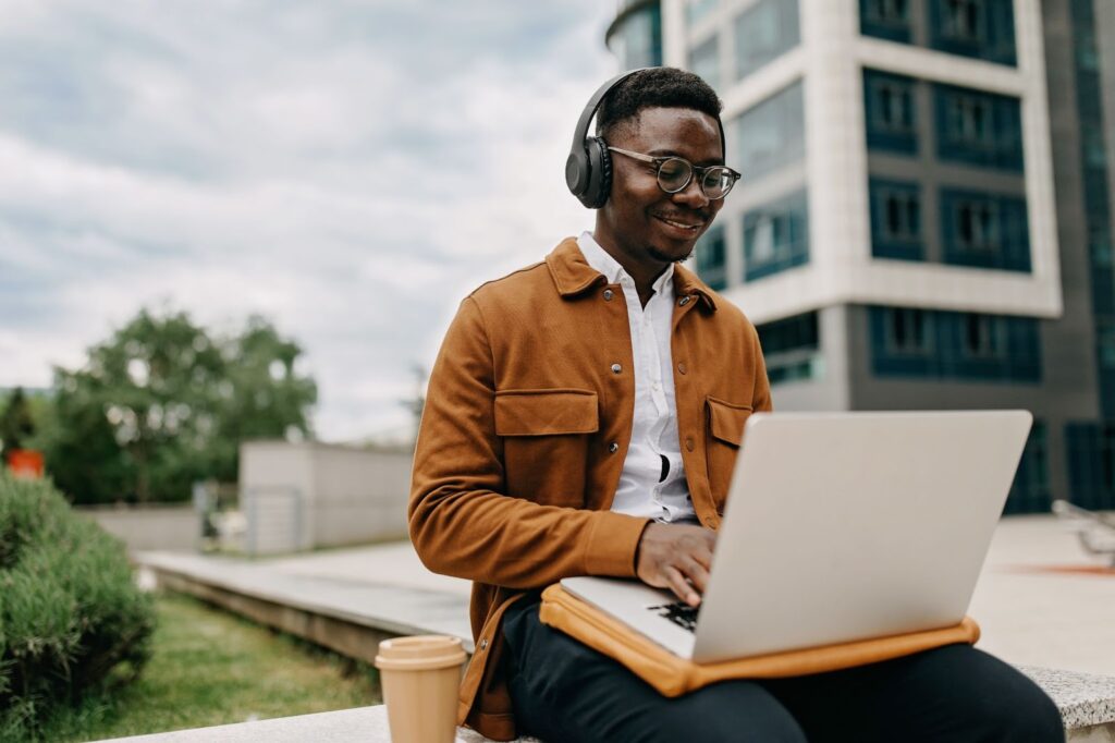 A Black male computer science student wearing glasses and headphones sits outside on a university campus and works on a laptop.