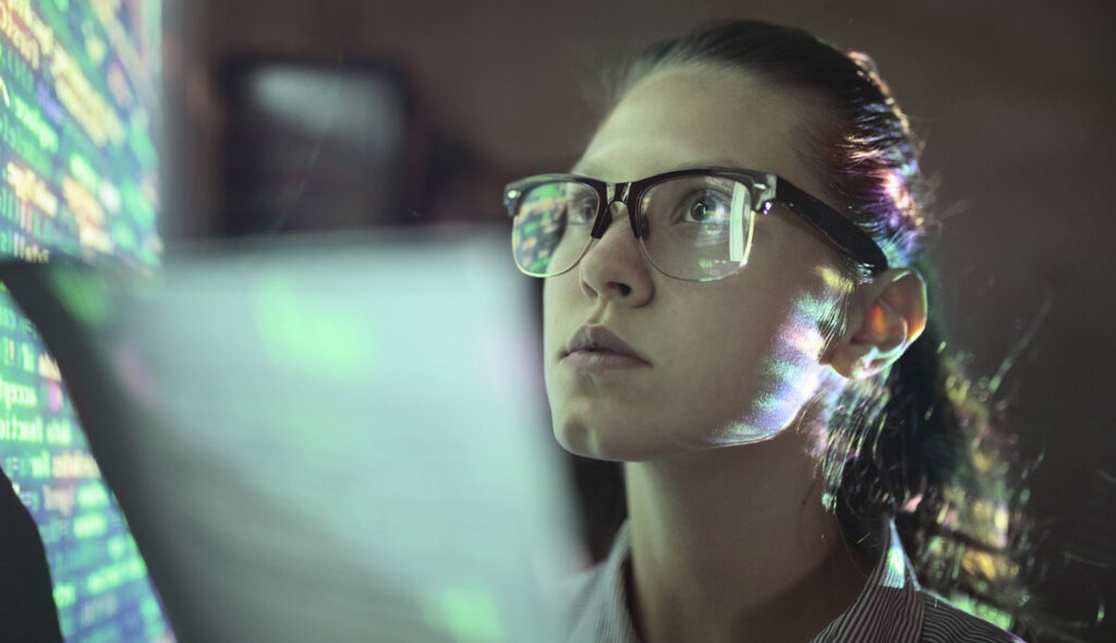 A computer scientist studies a screen of code that reflects in the lenses of her glasses.