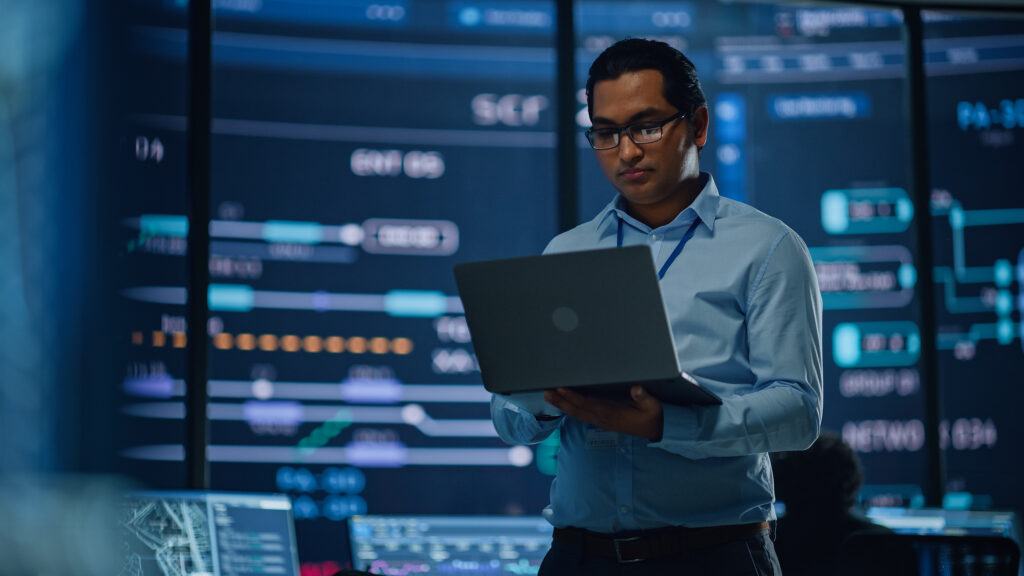A computer scientist studies a laptop while standing in a server room.